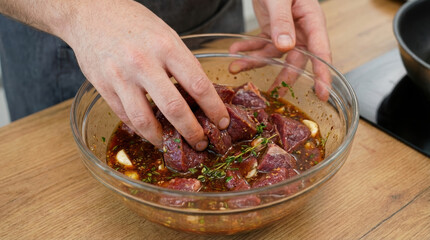 Marinated beef chunks in glass bowl with herbs and garlic being mixed by hands, savory and aromatic preparation for cooking