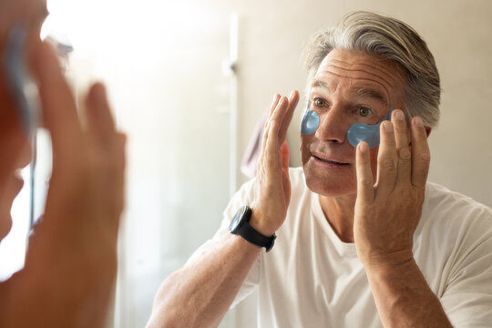 Middle-aged man applying undereye gel patches in home bathroom mirror wearing white tee black watch