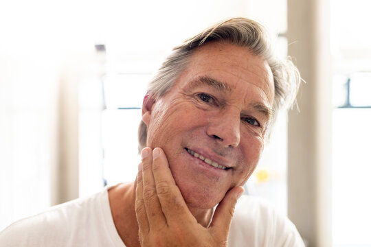 Senior man touching chin and making eye contact while wearing white T-shirt near backlit windows