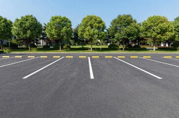 Empty Asphalt Parking Lot with White Stall Lines and Green Trees Under a Clear Blue Sky