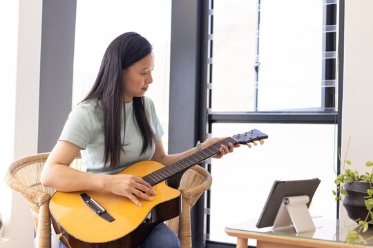 Asian woman sitting in woven rattan chair playing acoustic guitar while checking tablet on stand