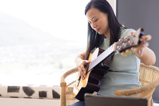 Asian woman sitting in rattan chair at home in top, jeans, adjusting acoustic guitar, copy space