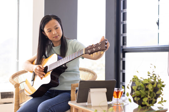 Asian woman sitting in chair at home tuning acoustic guitar by windows with tablet, green top