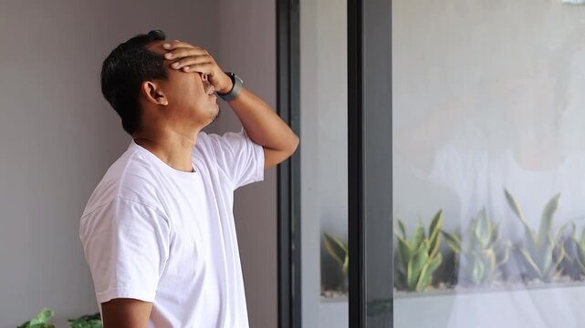 Frustrated Asian man standing near the window while thinking and scratching his head