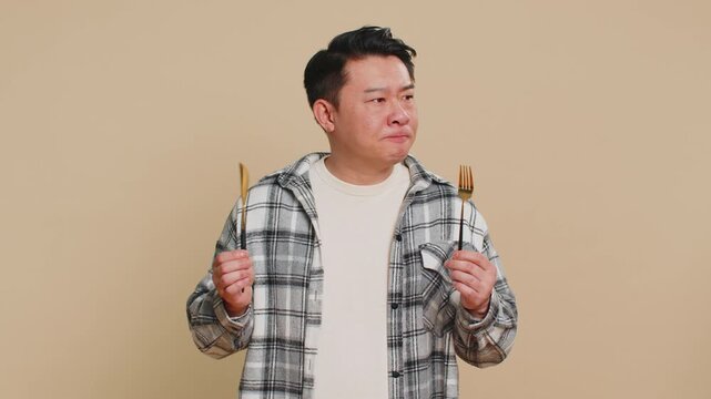 Ready to eat. Hungry Asian Chinese middle-aged man waiting for serving dinner dishes with with restlessness holding cutlery fork knife, will appreciate delicious restaurant meal on beige background