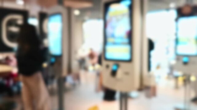 Unfocused footage of a modern fast-food restaurant interior. People use self-service digital kiosks with colorful food menus to order.