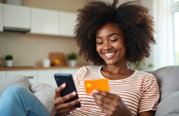 Naklejka premium Smiling young Black woman holds credit card and phone. She enters card number for online shopping purchase, paying bills from home couch. Happy consumer buys goods digitally.