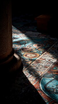In a softly illuminated room, a vibrant, intricately woven rug catches the light, while a prominent column stands in the foreground, adding depth to the scene
