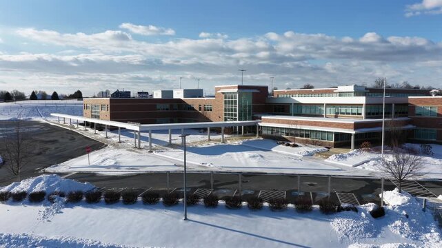 Aerial view of snow-covered school campus in Manheim, Pennsylvania, showing modern buildings, empty parking areas and winter landscape under blue skies. Quiet suburban American winter scene.