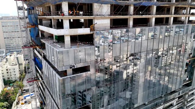 Close aerial of a high-rise under construction glass fa&ccedil;ade rising below active concrete floors, scaffolding, and workers shaping the skyline of a fast-growing urban business district.