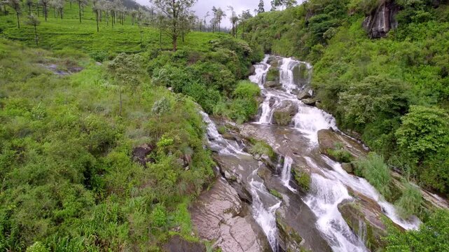 Cascading Ramboda waterfall moves across rocky green hillside forest in Nuwara Eliya Sri Lanka, aerial approach