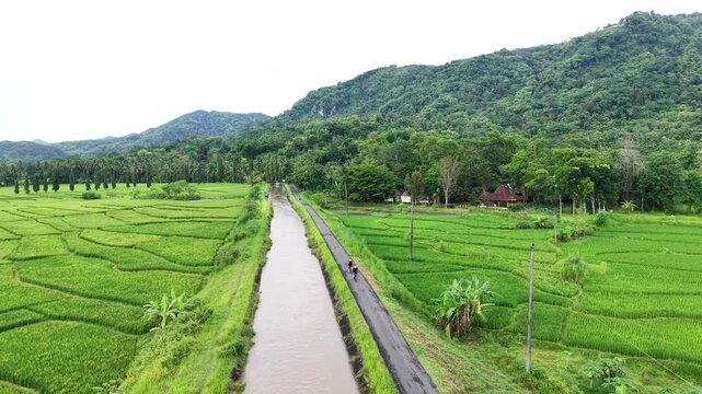 Aerial view of a couple cycling through the countryside along the Mataram river in Yogyakarta and the expanse of the Nanggulan rice fields. Cinematic footage, nature sports.
