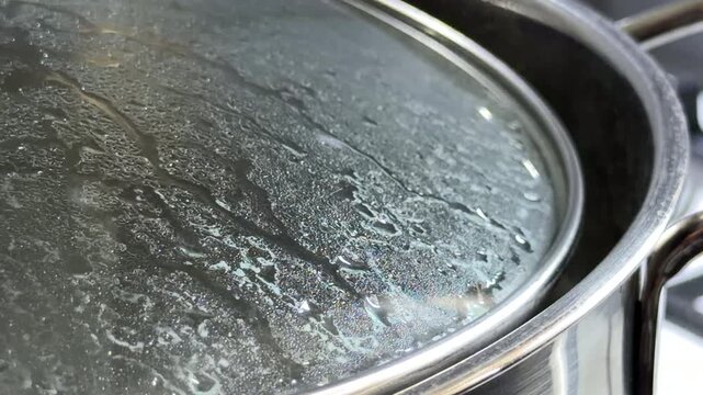 Water boils intensely inside a stainless steel pot covered with a glass lid. The trapped steam condenses into droplets as the water gurgles and splashes against the surface.