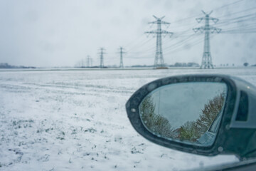 Fototapeta premium Winter landscape with snow covered field and power lines seen from car side mirror