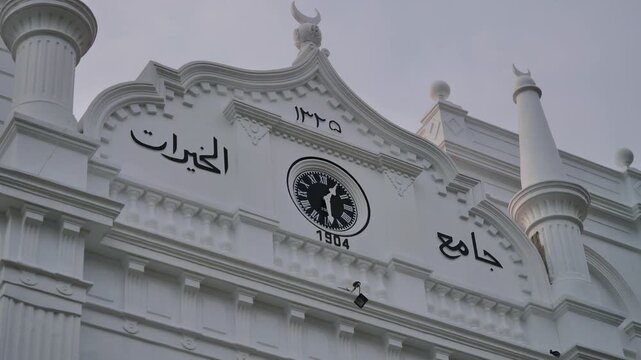 Establishing view showcasing historic white Islamic mosque facade, featuring detailed architectural molding, traditional crescent finials, central vintage 1904 analog clock, and Arabic calligraphy aga