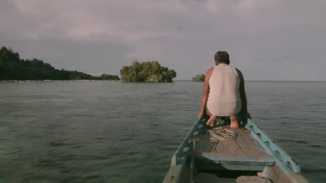 Local sailor sitting on the bow of a rustic wooden canoe sailing in the Togean Islands, Indonesia. Afternoon sunlight and calm water create a tropical paradise atmosphere.