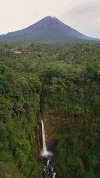 Vertical drone footage of Kapas Biru Waterfall surrounded by dense tropical rainforest with fresh water cascading into a rocky river while Mount Semeru active volcano appears visible in the background