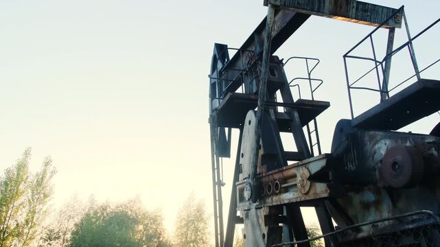 Rusty pumpjack tower fills frame with rivets showing wear by ladder. Horsehead lowers while counterweight spins near gearbox driving cycle