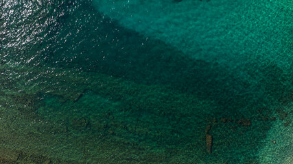 Top-down aerial shot of the Mediterranean Sea. The crystal-clear turquoise water reveals the intricate patterns of a dark, rocky seabed beneath the surface, creating a shimmering texture on the water.