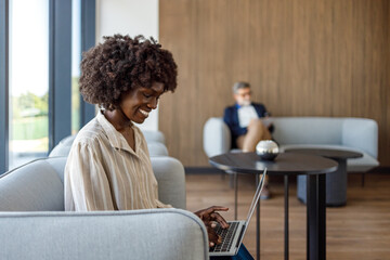 Confident businesswoman with afro hairstyle working over laptop while relaxing on sofa in modern...