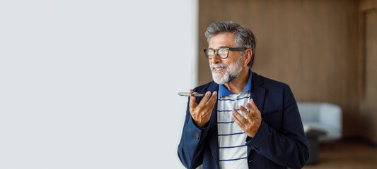 Confident mature businessman in eyeglasses using mobile phone to send voice message while standing...