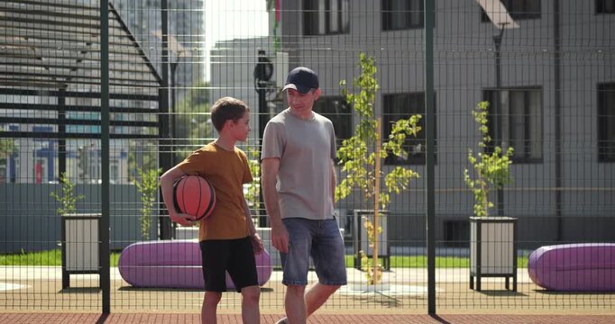 Father walks with son on outdoor court and talks with confidence. Confidence moment, support and positive example. Happy boy holds ball and gains confidence from parent. Setting child up for future.