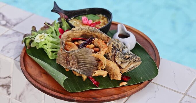 Crispy fried whole fish served on a banana leaf and wooden tray by a sparkling blue swimming pool. A concept of luxury dining, exotic cuisine, tropical vacation, gourmet seafood, and resort lifestyle.
