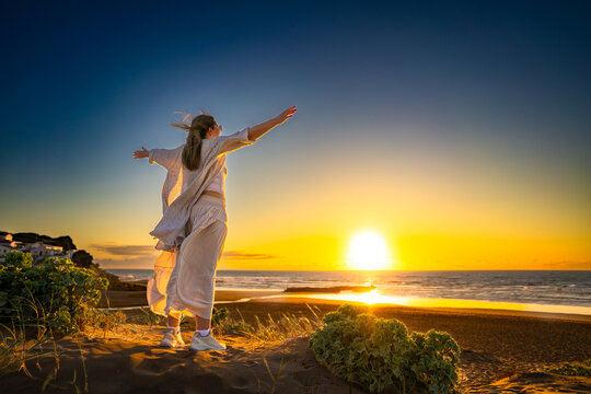 Beautiful middle-aged woman standing with outstretched arms on sandy beach on sunset on windy spring day. Back view. Monte Clerigo beach on Algarve coast in Portugal