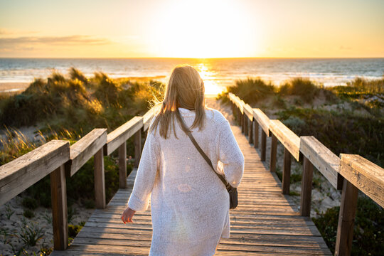 Beautiful middle-aged woman tourist walking down stairs overlooking sandy beach on sunset in spring. Back view	
