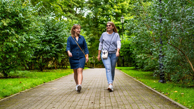Two happy middle-aged women walking in city park on spring day. Meeting of friends in park. Front view	