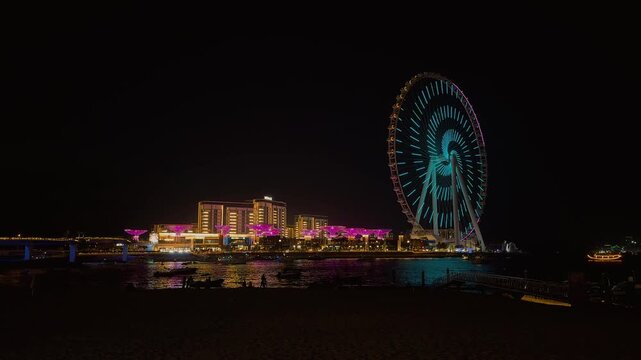 Night view of people walking on the pedestrian bridge towards the illuminated Ain Dubai Ferris wheel. The iconic landmark changes colors while city lights glow across the water.

