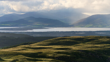 Hillsides glow under the soft light. Mountains stand silhouetted in the distance. Forests fill the valleys below the hills. Clouds soften the sky above mountain peaks.