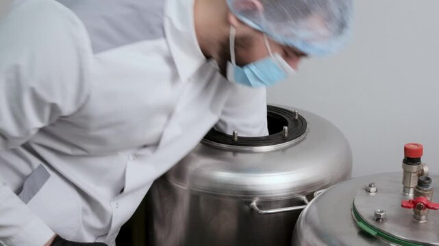 Laboratory worker in white coat opens a stainless steel autoclave, removes a sterilized grain jar and secures the lid again. Clean sterile substrate processing under controlled laboratory conditions.
