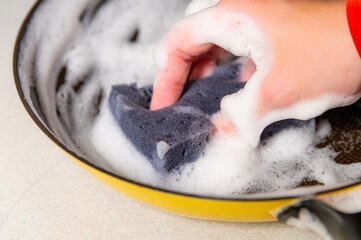 Obraz premium Hand scrubbing a soapy sponge in a yellow frying pan filled with bubbles on a light countertop, showcasing kitchen cleaning activity and dishwashing essentials