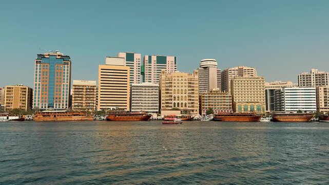 Scenic panning shot of Dubai Creek featuring the Deira skyline with modern skyscrapers and traditional wooden dhow boats docked along the sunny waterfront in United Arab Emirates.

