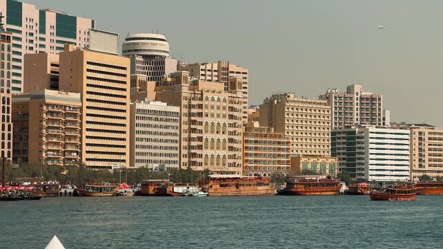 Scenic panning shot of Dubai Creek featuring the Deira skyline with modern skyscrapers and traditional wooden dhow boats docked along the sunny waterfront in United Arab Emirates.

