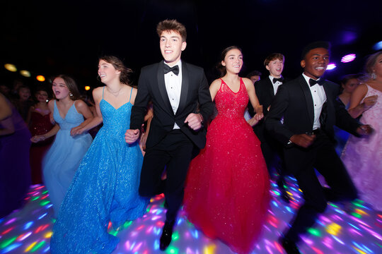Group of teenagers in formal prom attire dancing together on a colorful lighted dance floor, wearing elegant gowns and tuxedos during a festive celebration