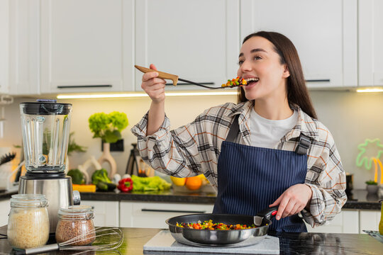 Young woman samples vegetables from frying pan while cooking in home kitchen for diet plan and daily wellness. Girl looks satisfied after taste test ready to serve homemade lunch in apartment indoors.