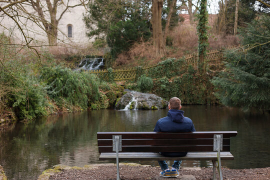 A man sits alone on a wooden bench facing a calm pond surrounded by trees and greenery. The tranquil setting with soft natural light creates a reflective and peaceful atmosphere. 