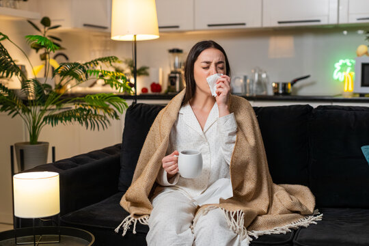 Young woman at night home under blanket with hot tea and medicine sips for sore throat cough. Caucasian girl on sofa at evening uses tissue sneezes feels fever fatigue so rests quiet asks help softly.