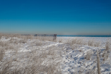 A winter scene shows a snow-covered beach with dry grass and a wooden fence in the foreground