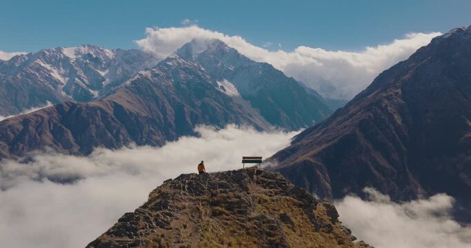 A traveler walking to a bench and sitting on top of a mountain peak above clouds in North Ossetia, Russia. Cinematic aerial view of a hiker enjoying Caucasian mountain landscape.