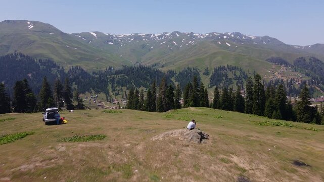 Aerial pan over Svaneti, Georgia shows village along a shallow river, a parked car, two people, dirt tracks, wooden houses, spruce slopes, snow ridges, and summer sky.