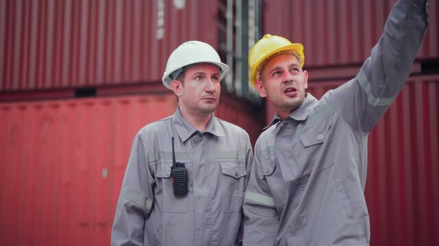 Two logistics workers in hard hats having a serious discussion over documents at a container port. Industrial teamwork and supply chain management concept.