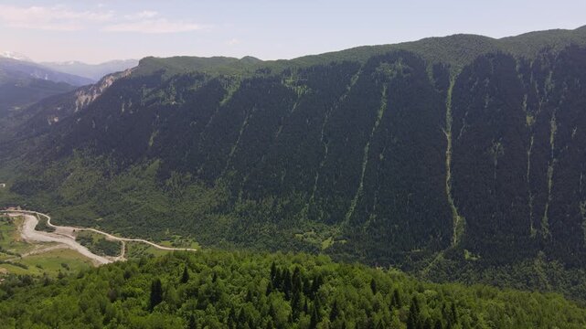 Aerial view shows Svaneti valley in Georgia as a braided pale river winds past meadows and hamlets toward Mount Ushba, with conifer cliffs, pastures, and erosion gullies.