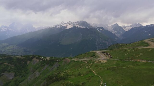 Aerial drone sweeps over alpine meadows and a dusty switchback road in Svaneti, Georgia. Snow streaked peaks rise, tiny hikers and pylons appear under soft, moody clouds.
