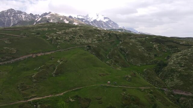 Aerial view of green pastures, ridgelines, and a winding dirt track near Mount Ushba in Svaneti, Georgia. A red shelter, hikers, and vehicles appear small in soft light.