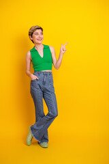 Young woman in green top and jeans stands against bright yellow background pointing with a smile © deagreez