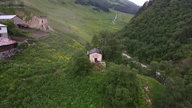 Aerial view shows stone chapel on grassy knoll, river flowing, Svan towers and houses near Mestia and Ushguli in Georgia, tiny figures, summer pastures, soft overcast light