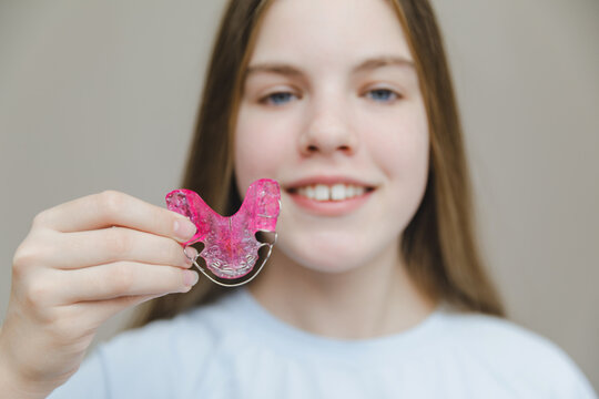 Smiling teenage girl holding pink removable orthodontic plate in front of her face, pediatric dentistry and professional bite correction concept